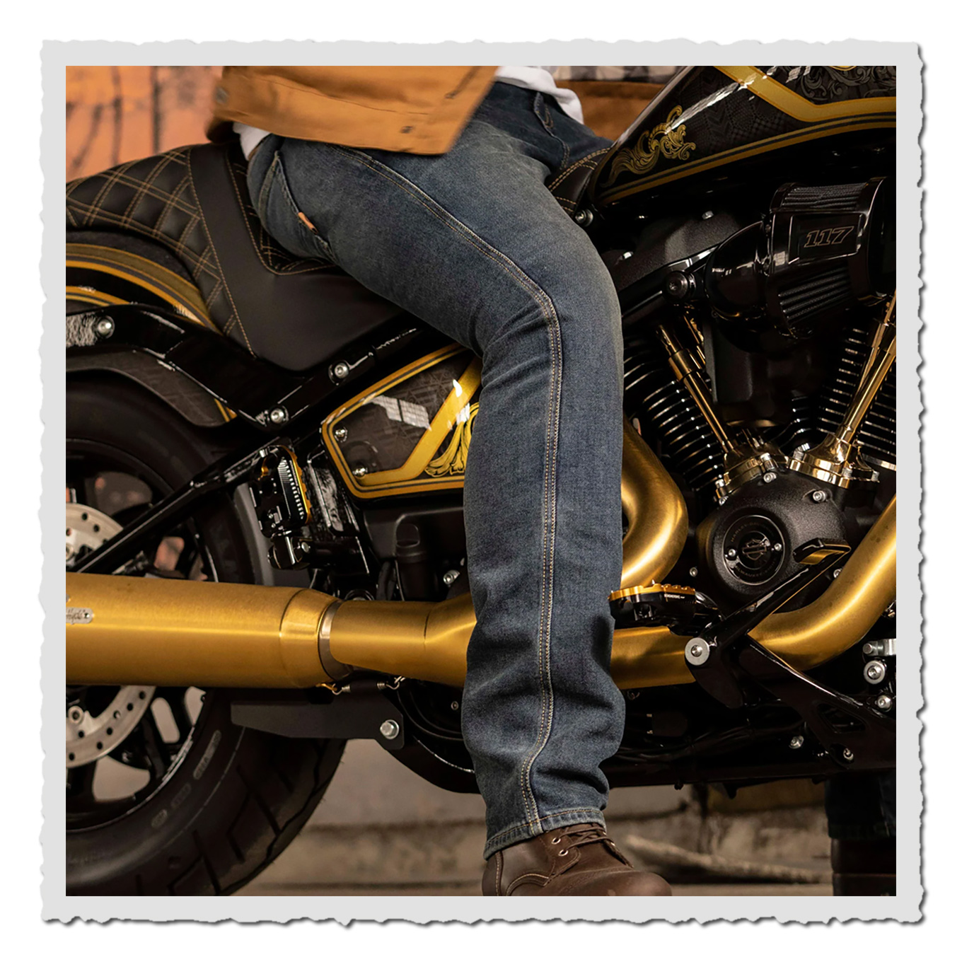 Nahaufnahme einer sitzenden Person in blauer Jeans neben Motor, Auspuff und schwarzem Motorradsitz.